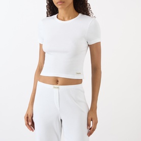 Model wearing Sporty and Rich Signature Logo Rib Baby T-Shirt in white, ribbed cotton, cropped fit, round neck, paired with white trousers.
