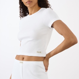 Model wearing Sporty and Rich Signature Logo Rib Baby T-Shirt in white, ribbed texture, round neck, short sleeves, styled with white skirt.