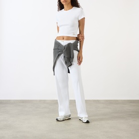 Model wearing Sporty and Rich Signature Logo Rib Baby T-Shirt in white, styled with white wide-leg trousers and grey shirt tied at waist.
