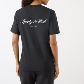 Model wearing Sporty and Rich Signature NY Logo T-Shirt in Faded Black, back view showing cursive logo print, paired with black trousers.