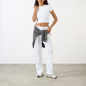 Model wearing Sporty and Rich Signature Logo Tank Top in white with white wide-leg trousers and grey jacket tied at waist.