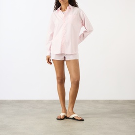 Model wearing Sporty and Rich Signature Logo Shirt in Clear Pink cotton, styled with white shorts and flat sandals