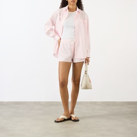Model wearing Sporty and Rich Signature Logo Shirt in Clear Pink with matching shorts, white tee, tan sandals, and cream handbag.