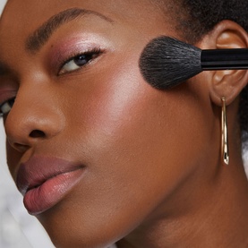 Model with Bobbi Brown brush applying powder to cheekbone, gold hoop earring visible, close-up beauty shot
