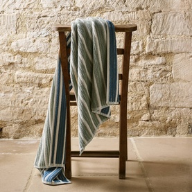 Christys bath towel in multi blue and white stripes, draped over a wooden chair against a stone wall backdrop.