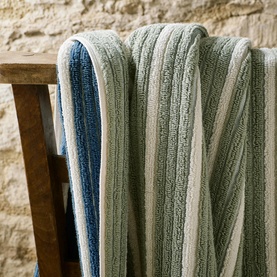 Christys bath towelling draped over a wooden chair, featuring ribbed stripe texture in soft sage green and blue multi tones