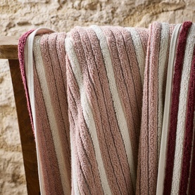 Christys bath towel in multi-stripe pink, rose and cream draped over a wooden chair against a stone wall background.