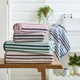 Deyongs Linear Stripe Towel in green, stacked with pink and grey striped towels on a wooden bench in a bathroom setting.