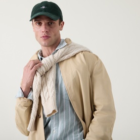Model with Gant Shield Logo Corduroy Cap in Storm Green, styled with a beige trench coat, cable-knit scarf and striped shirt.