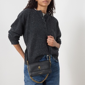 Model carrying Kurt Geiger Sloane Crossbody Bag in black smooth leather with gold-tone chain strap and eagle head hardware, styled with grey