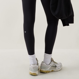 Model wearing Lululemon black leggings, shown from behind, styled with white ankle socks and grey trainers, holding a dark sweatshirt.