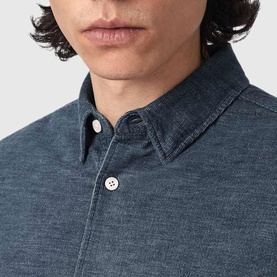 Model wearing All Saints shirt in Moonstone Blue, close-up of button-down collar and chest buttons on chambray-style fabric.