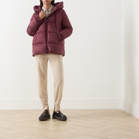 Model wearing a burgundy hooded puffer jacket, styled with cream tailored trousers and black loafers