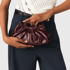 Model carrying a small burgundy ruched leather clutch bag with gold top handle, styled with navy trousers and white top