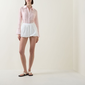 Model wearing Aligne Paulo Bubble Shorts in white poplin with bubble hem, styled with a pink shirt and flat sandals.