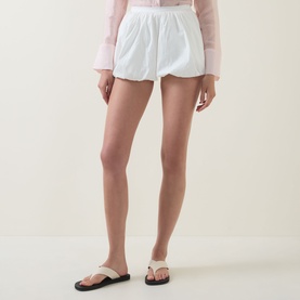 Model wearing Aligne Paulo Bubble Shorts in white with bubble hem, styled with black flip-flops and a pink long-sleeve top.