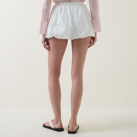 Model wearing Aligne Paulo Bubble Shorts in white with bubble hem, styled with a pink top and black flat sandals.