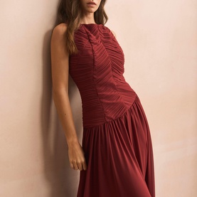 Model wearing a dark red sleeveless maxi dress with ruched bodice and flowing skirt, leaning against a neutral wall