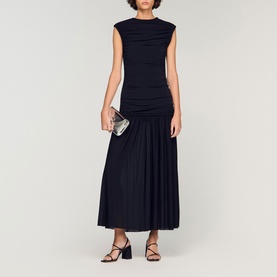 Model wearing Sandro Womens French Dress in black, ruched bodice, pleated maxi skirt, styled with black heeled sandals and silver clutch bag.