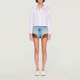Model wearing Sandro white embroidered blouse and blue denim shorts with brown suede trim, styled with white trainers.