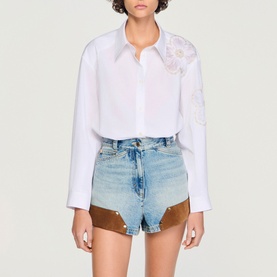 Model wearing Sandro white shirt with floral lace detail, styled with light blue denim shorts with tan panel trim.