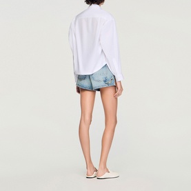 Model wearing Sandro white oversized shirt with light blue denim mini shorts and white slip-on shoes, back view