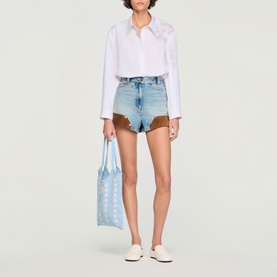 Model wearing Sandro white shirt with floral collar detail and light blue denim shorts, carrying a light blue tote bag, styled with white shoes.