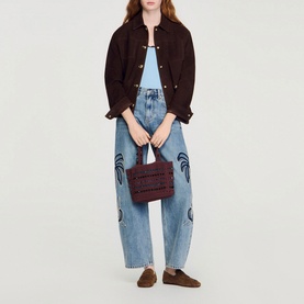 Model carrying Sandro brown quilted tote bag, styled with dark brown suede jacket, light blue crop top, and printed wide-leg jeans