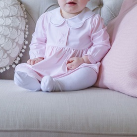 Baby wearing Emile Et Rose pink long-sleeve dress with white Peter Pan collar, styled with white tights and booties, seated on grey sofa.