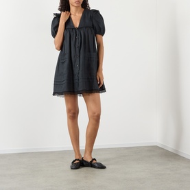 Model wearing Ganni black linen mini dress with puff sleeves, V-neck, and lace hem trim, styled with black flat mules