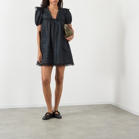 Model wearing Ganni black linen mini dress with puff sleeves, V-neckline, button front and lace hem trim, styled with black slides and olive mini bag.