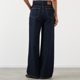 Model wearing Ganni Lari Wide-Leg Jeans in Rinse, showing rear view with tobacco stitching, logo patch, and five-pocket styling.