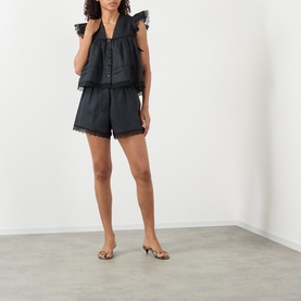 Model wearing Ganni Angel Linen Blouse in black with frill shoulders, lace trim, styled with matching black shorts and gold heeled sandals.