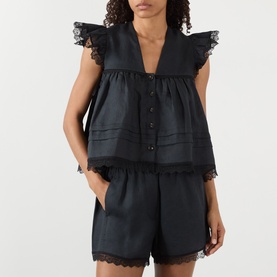 Model wearing Ganni Angel Linen Blouse in black with lace trim, frill shoulders, and button placket, styled with matching black linen shorts.