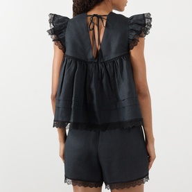 Model wearing Ganni Angel Linen Blouse in black, rear view showing tie fastening, lace trim, frill shoulders and layered pleats. Styled with black