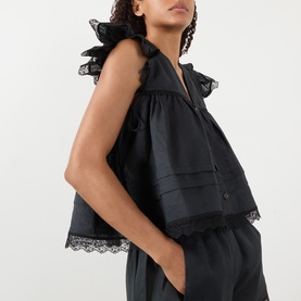 Model wearing Ganni Angel Linen Blouse in black with lace trim, frill shoulders, and V-neckline, styled with matching black wide-leg trousers