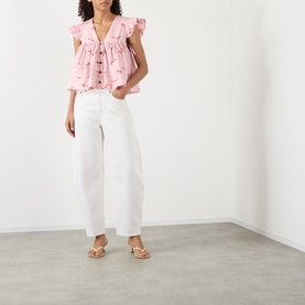 Model wearing Ganni pink cotton blouse with ruffle trim and button front, styled with white wide-leg jeans and nude heeled sandals