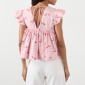 Model wearing Ganni pink cotton blouse with floral embroidery, flutter sleeves, deep V-back with tie detail, styled with white trousers