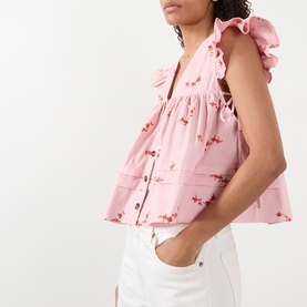 Model wearing Ganni pink floral embroidered cotton blouse with ruffle shoulders and button front, styled with white trousers