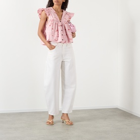 Model wearing Ganni pink cotton blouse with frill shoulders and floral detail, styled with white wide-leg jeans and gold strappy heels