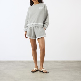 Model wearing Ganni Paloma shorts in grey cotton with frayed hem edges, styled with matching sweatshirt and tan sandals.