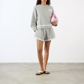 Model wearing Ganni Paloma shorts in grey cotton with frayed hem trim, styled with matching sweatshirt and brown sandals, carrying pink mini bag