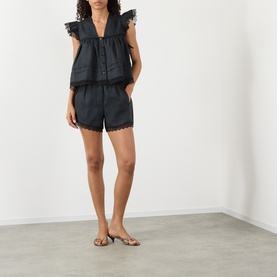 Model wearing Ganni black linen shorts with scalloped broderie anglaise hem, styled with matching black ruffle top and gold heeled sandals