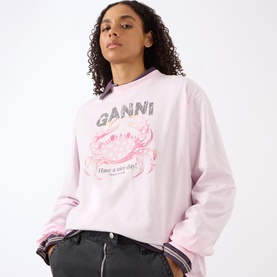 Model wearing Ganni Chalk Pink cotton sweatshirt with crab graphic print, styled with dark grey trousers and layered striped hem detail.