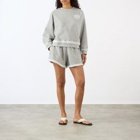 Model wearing Ganni Isoli Logo Lace-Trim Sweatshirt in Paloma with matching grey shorts and white sandals.