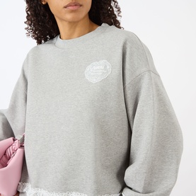 Model wearing Ganni Isoli Logo Lace-Trim Sweatshirt in Paloma grey with chest logo patch and lace-trim hem, styled with a pink bag.