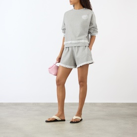 Model wearing Ganni Isoli Logo Lace-Trim Sweatshirt in Paloma with matching grey shorts, styled with white slides and a pink mini bag.
