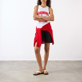 Model wearing Ganni Cherry Logo Ribbed Tank Top in Bright White, styled with black cycling shorts, red shirt tied at waist and sandals.