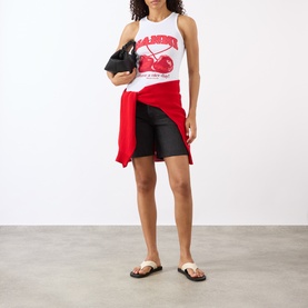 Model wearing Ganni Cherry Logo Ribbed Tank Top in Bright White with black shorts, red shirt tied at waist, and black mini bag.