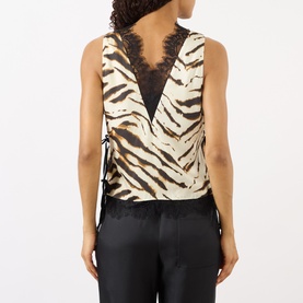 Model wearing Ganni Zebra Print Lace-Trim Blouse in Egret, showing V-shaped back, lace trim, side-tie fastenings, styled with black satin trousers.